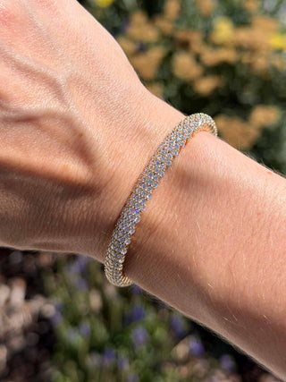 Close-up of a woman's wrist wearing a pavé-set stretch tennis bracelet in yellow gold, outdoors with natural sunlight and blurred flowers in the background.