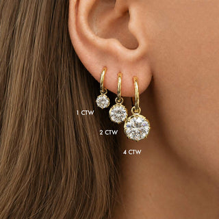 Close-up of a woman's ear wearing three gold huggie hoop earrings with roundlab grown diamonds in a georgian setting in different sizes, labeled 1 CTW, 2 CTW, and 4 CTW.