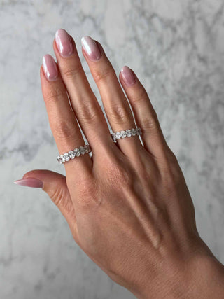 A close-up of a woman's hand with pink manicured nails wearing two 14k white gold oval lab grown diamond eternity bands, photographed against a marble background.