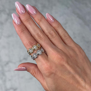 A close-up of a hand with glossy pink manicured nails wearing two stacked eternity bands with puffy bezel setting and round lab grown diamonds in yellow gold and white gold, set against a soft gray marble background.