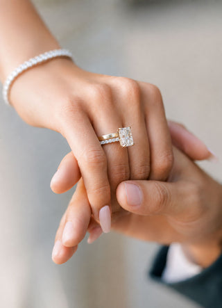 A close-up shot of a bride's hand wearing a gold band and a radiant cut engagement ring with pave band, her hand held by a man's hand.