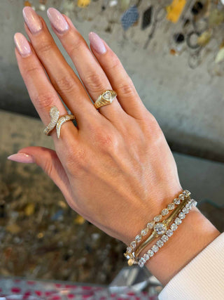 A close-up of a woman's hand wearing multiple gold and diamond jewelry pieces, including a snake-shaped ring, a heart-shaped diamond fluted ring, and three stacked diamond bracelets, against a blurred background.