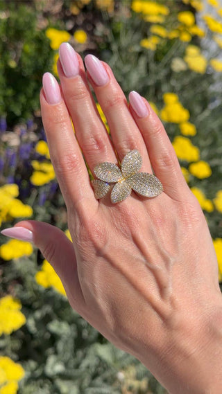 Video of Lab Grown Diamond flower ring shown on a hand in natural sunlight, outdoors with blurred flowers in background.